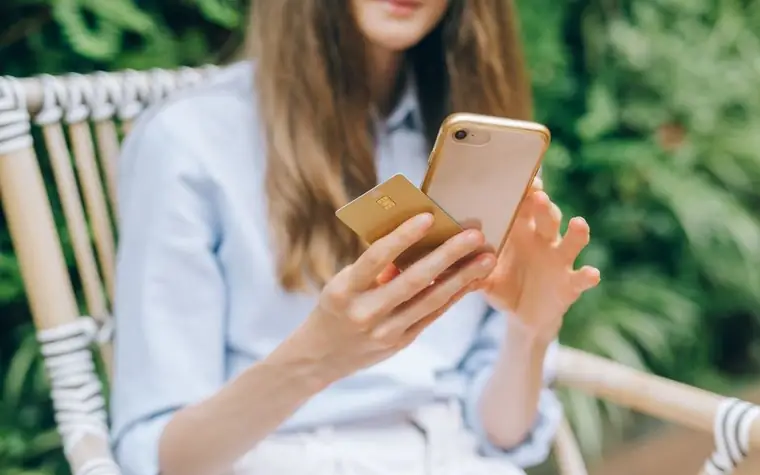 카페 야외 의자에 앉아 금색 카드를 들고 스마트폰을 보는 여성
